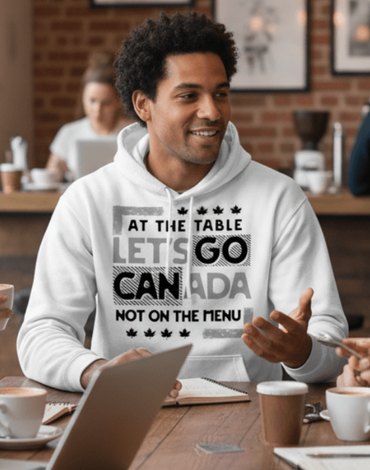Man wearing a white hoodie with text design, At the table, Let's go Canada, Not on the menu, seated indoors.
