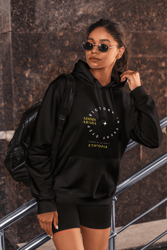 Woman wearing a black Ethiopia Addis Ababa hoodie with coordinates text and graphics, standing against a textured wall.