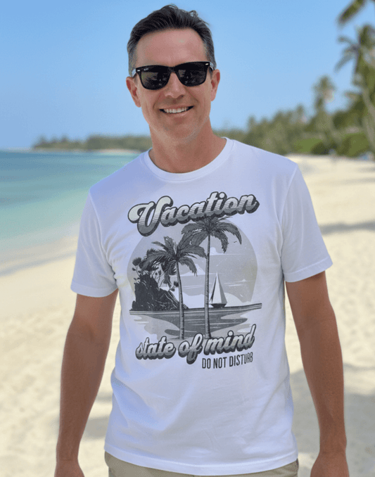 Man standing on the beach wearing a white t-shirt with a graphic print and text that says Vacation - State of Mind - Do not disturb