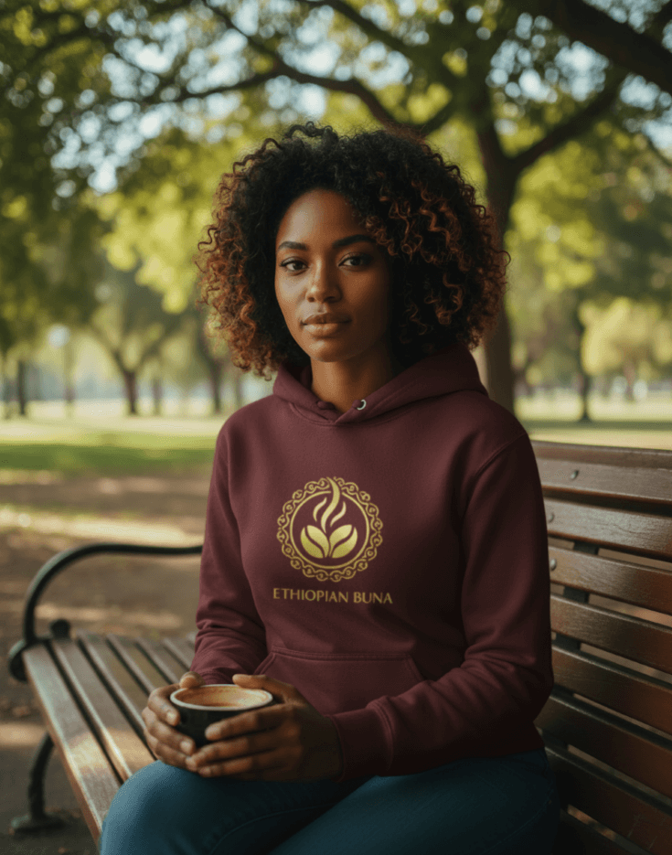 Woman wearing a maroon Ethiopian Buna graphic print hoodie holding a cup of coffee in a cozy setting