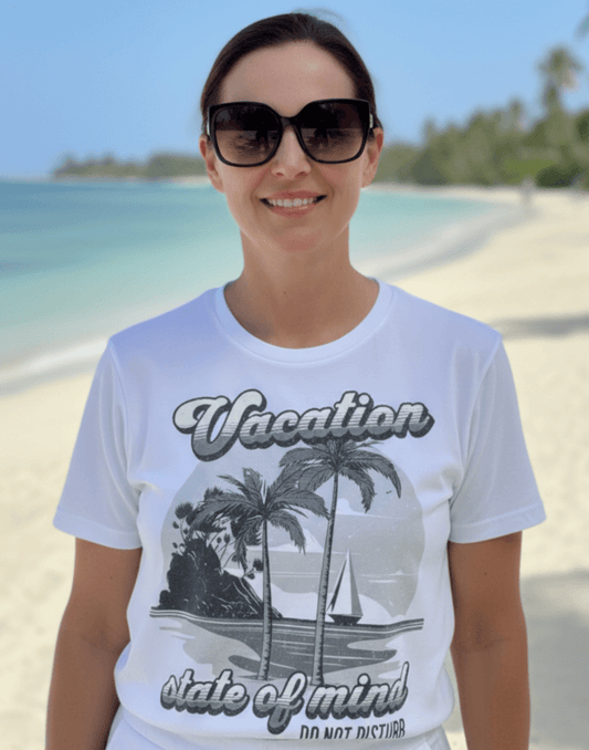 Woman standing on the beach wearing a white t-shirt with a graphic print and text that says Vacation - State of Mind - Do not disturb