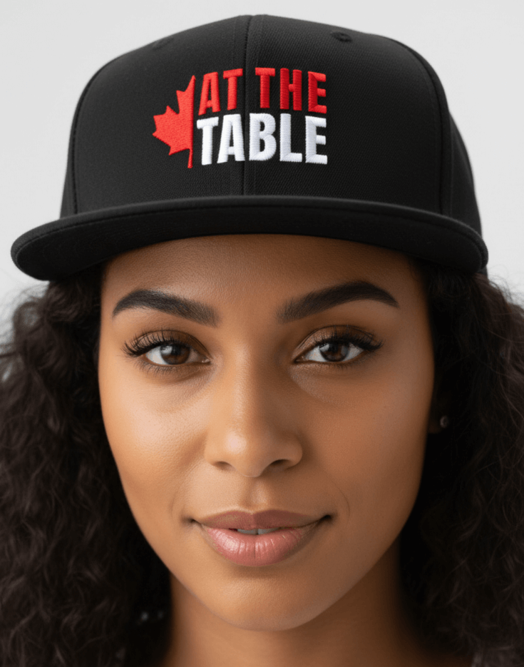 Woman wearing a black flat billed snap back cap with 'At the Table' logo, blurred background
