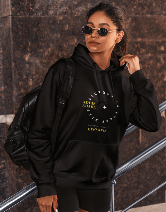 Woman wearing a black Ethiopia Addis Ababa hoodie with coordinates text and graphics, standing against a textured wall.