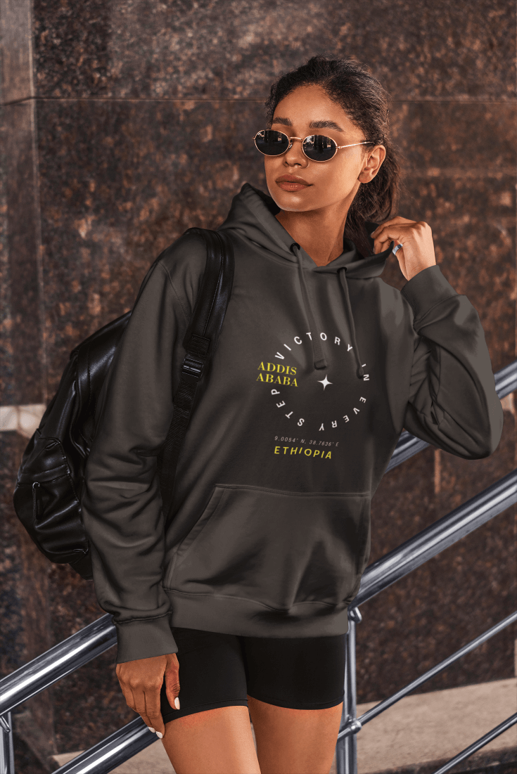 A woman wearing a dark heather gray Ethiopia Addis Ababa hoodie with coordinates text and graphics next to a textured wall