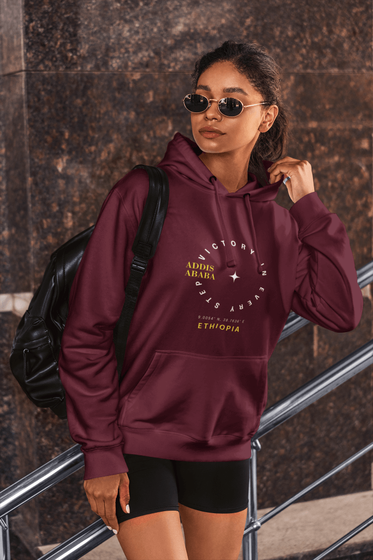 A woman wearing a maroon red Ethiopia Addis Ababa hoodie with coordinates text and graphics next to a textured wall