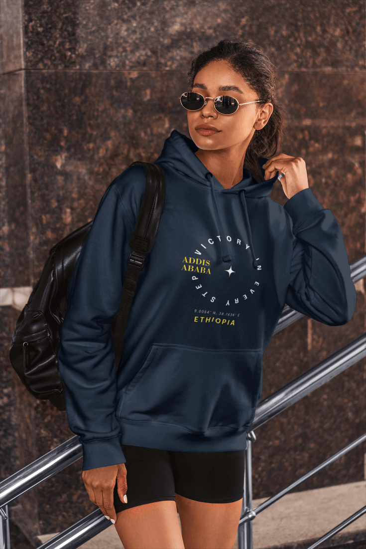 A woman wearing a navy blue Ethiopia Addis Ababa hoodie with coordinates text and graphics next to a textured wall