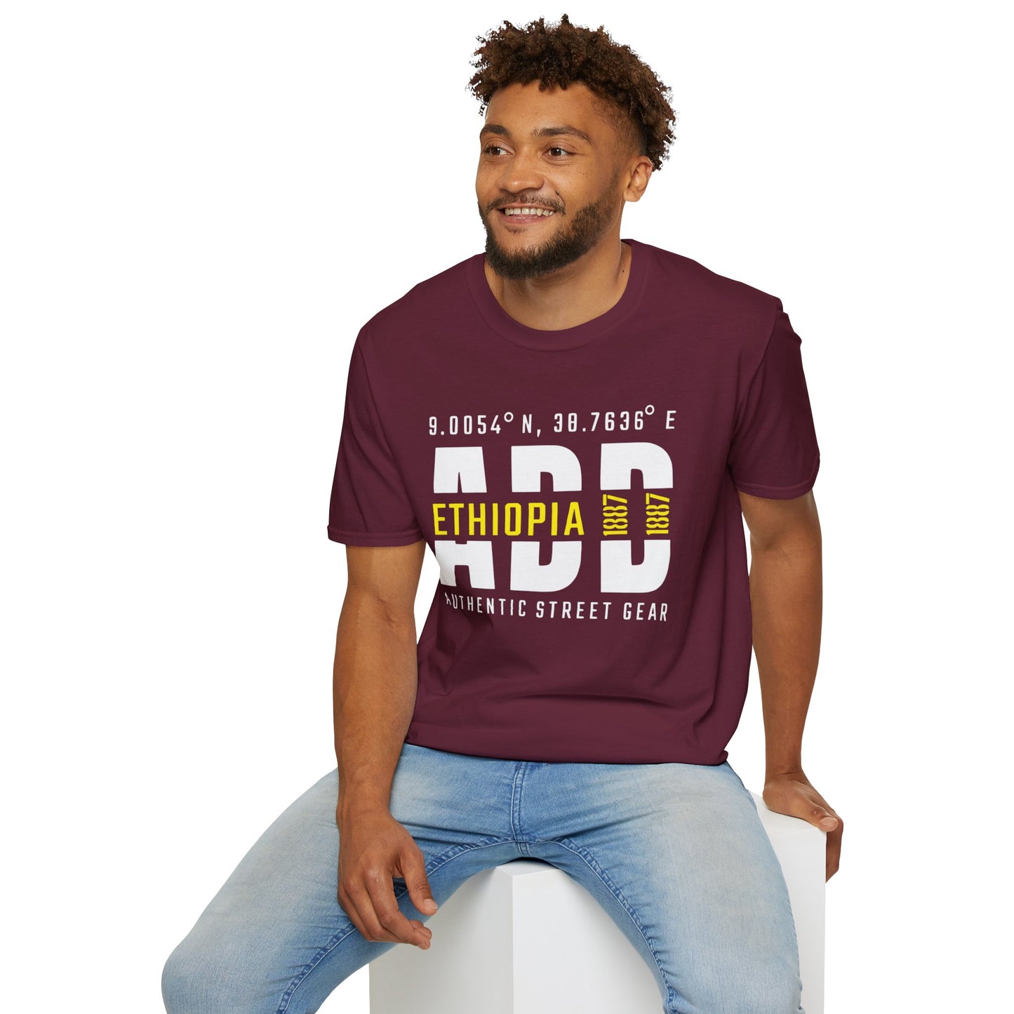 Man wearing a maroon t-shirt with Addis Ababa, Ethiopia, ADD Airport code and coordinates, sitting on a white stool against a white background.
